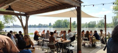Une guinguette au bord de l’eau, tables occupées sous la voile tendue.